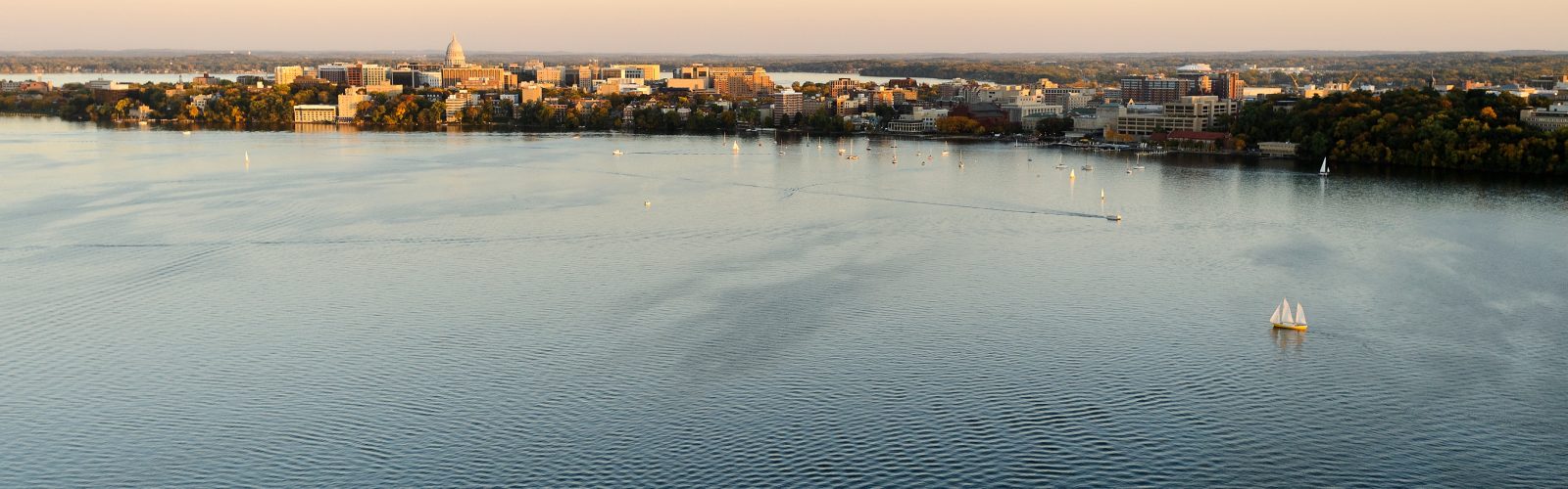 The Wisconsin State Capitol dots the downtown Madison skyline, at left, and the University of Wisconsin-Madison campus, at right, wraps along the Lake Mendota shoreline in an aerial view during an autumn sunset on Oct. 5, 2011. In the foreground are sailboats on Lake Mendota. In the background is Lake Monona, The photograph was made from a helicopter looking southeast. (Photo by Jeff Miller/UW-Madison)