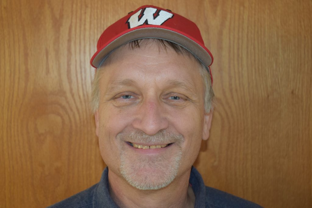 John Lyons wearing a red baseball hat with white W on front, standing in front of wood paneling and smiling