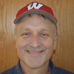 John Lyons wearing a red baseball hat with white W on front, standing in front of wood paneling and smiling