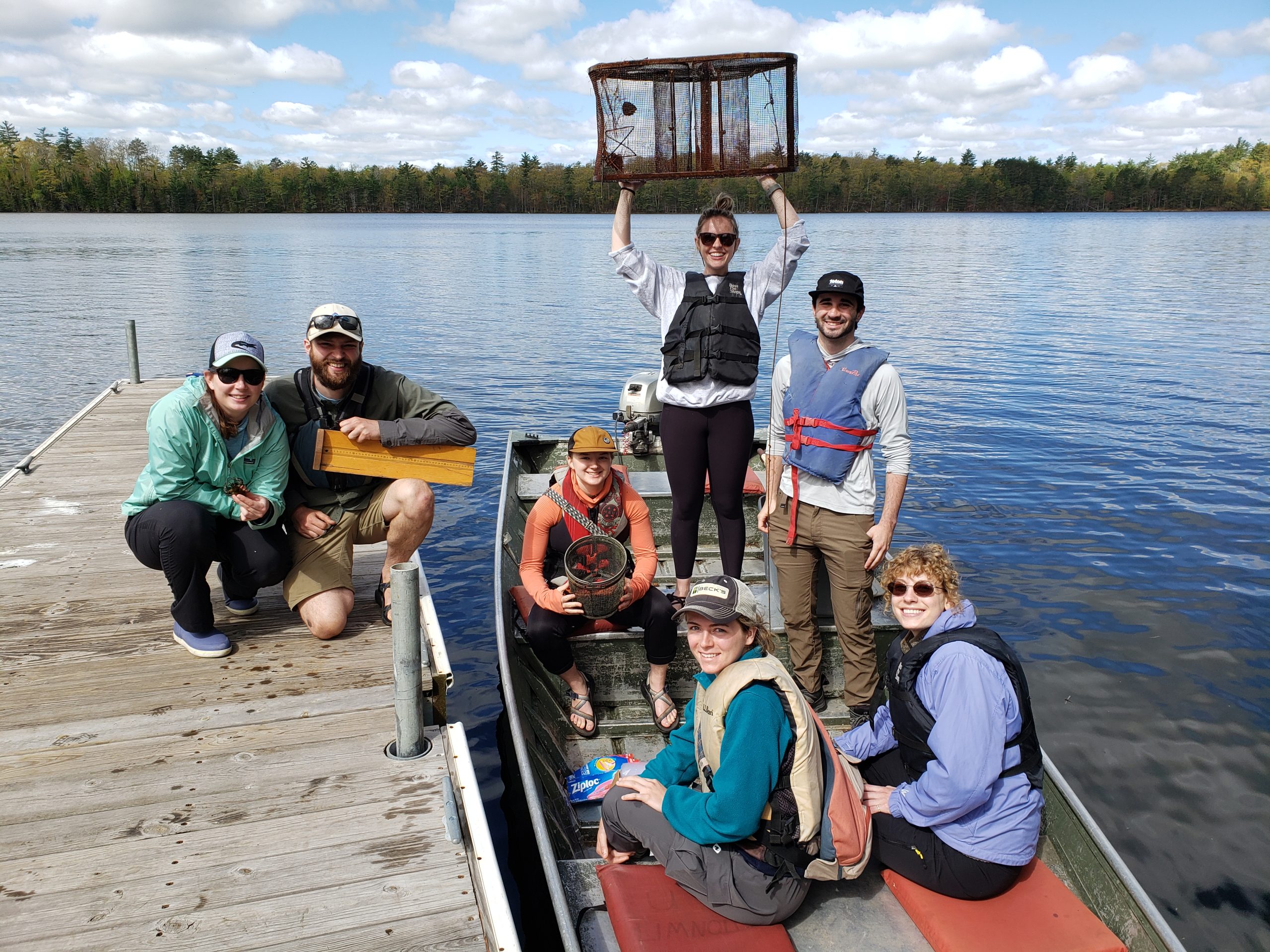Two people kneeling on dock, 5 people in boat beside boat dock, one holding trap over their head. All looking at the camera