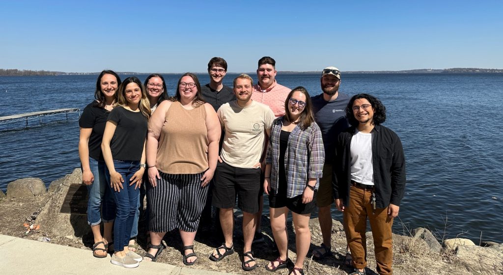 Group of CFL Grad students standing in front of Lake Mendota with bright sun shine and blue sky in background.