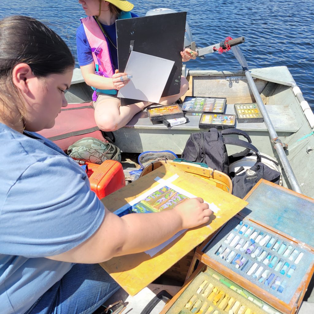 A couple of the undergraduate artist interns sits on dock on a sunny day on lake in Northern Wisconsin with art supplies around her and in her lap drawing and painting.